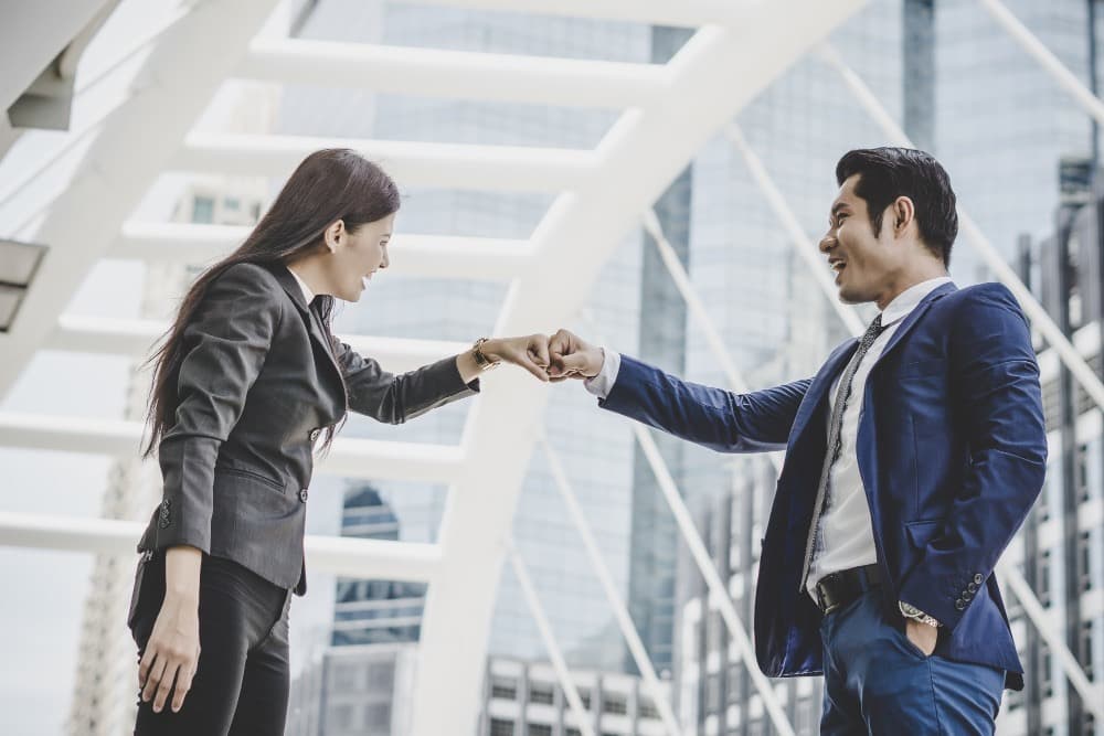 Two business professionals smiling and fist-bumping outdoors in a modern city setting.